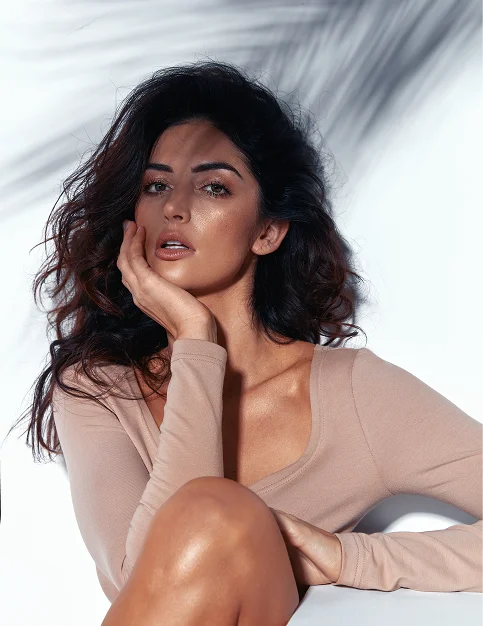 Woman in beige bodysuit posing with hand on face, dark wavy hair, professional fashion photography with soft lighting