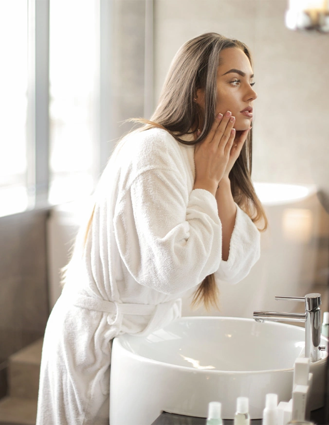 Woman in white bathrobe applying skincare product while looking in bathroom mirror by sink - Acne scars In Cleveland, OH