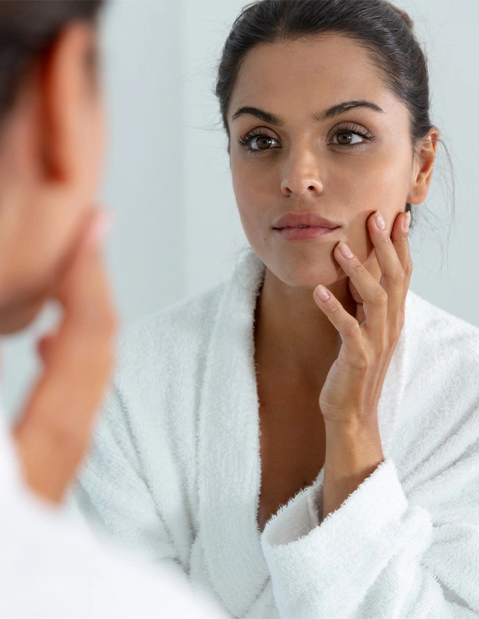 Woman examining her facial skin in mirror while wearing white robe for skincare routine - Dull skin In Cleveland, OH