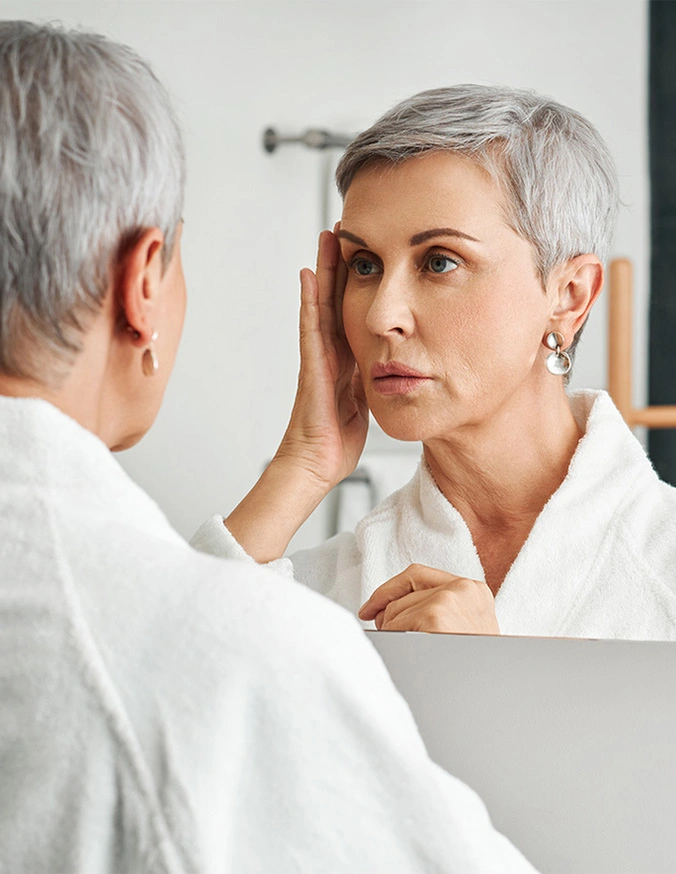 Woman with short gray hair examining her face and skin in bathroom mirror while touching cheek - Loose skin In Cleveland, OH